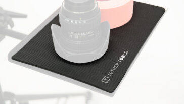 Close-up of a camera lens resting on a black textured anti-slip mat, with a pink roll in the background.