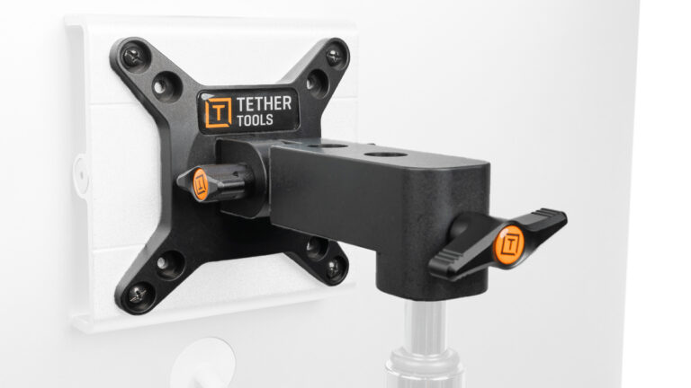 Black adjustable monitor arm attached to the back of a white monitor via a VESA plate, branded Tether Tools, with orange knobs for tightening