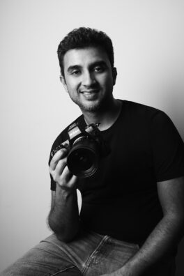 Black-and-white portrait of a man smiling and holding a Nikon camera at chest level in a studio setting.