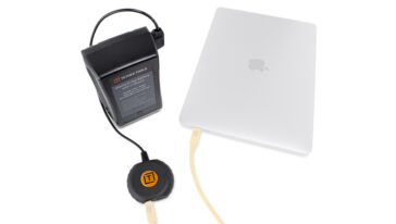 External battery pack connected to a white laptop via a power cable for portable charging. In the foreground is a round power puck on the cable.