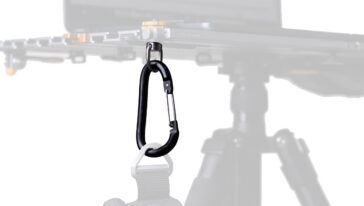 Close-up of a black carabiner clipped to a strap on a drone/rig, with a tripod blurred in the background.