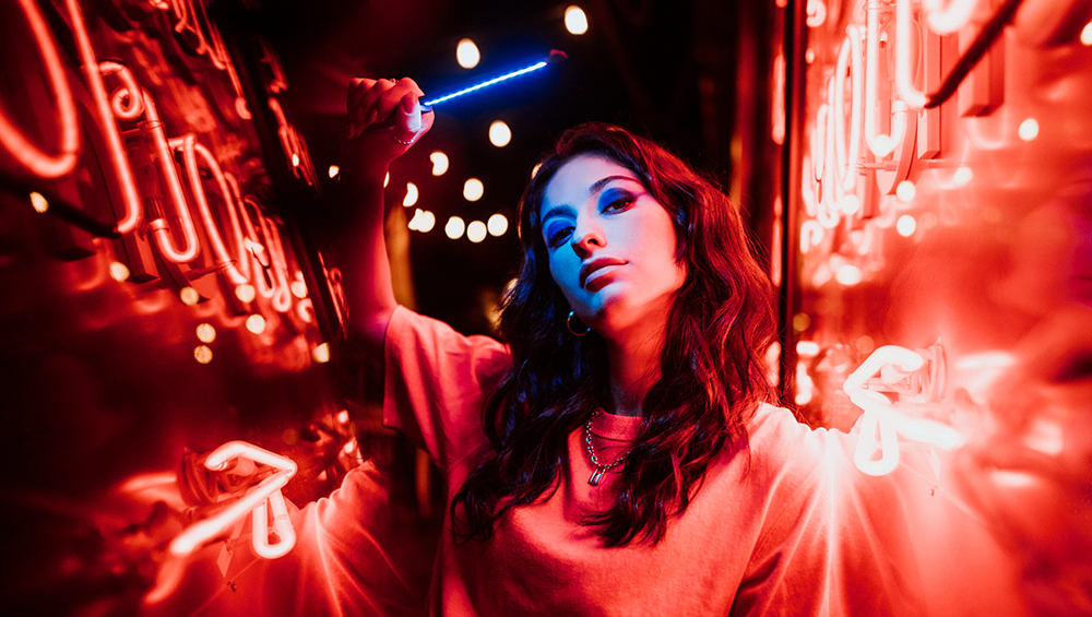 Woman with wavy dark hair in a pink hoodie stands in a red neon-lit tunnel, holding a blue glow stick alive with light.