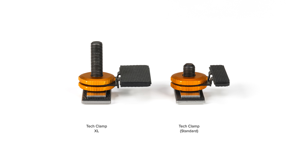 Two orange Tech Clamps on a white background: left is XL with a tall bolt and black pedal, right is Standard with a shorter bolt and pedal.