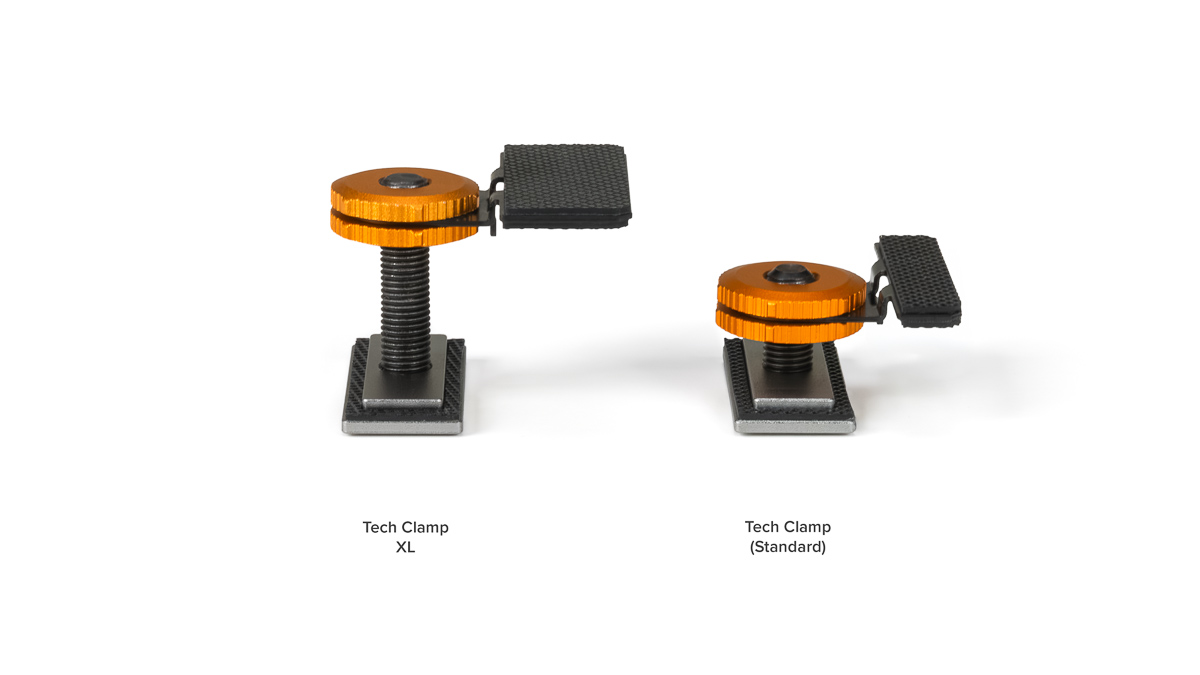 Two Tech Clamp devices—XL on the left and Standard on the right—orange adjustment wheels, black textured foot pedals, on square metal bases against a white background.