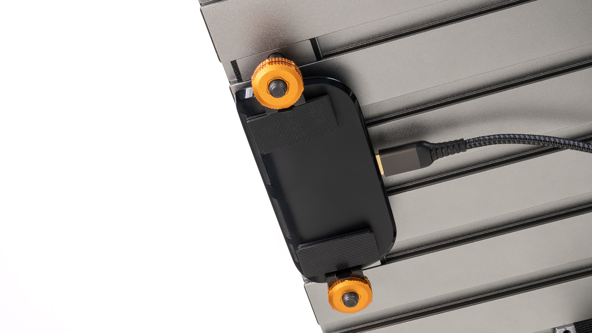 Close-up of a black rectangular device mounted on a metal slat, with orange knobs and a braided USB-C cable connected on the right side.