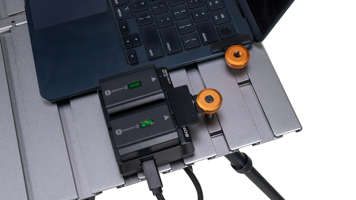 Laptop connected to a dual external battery charger on a metal surface with orange adjustment knobs, powering the device