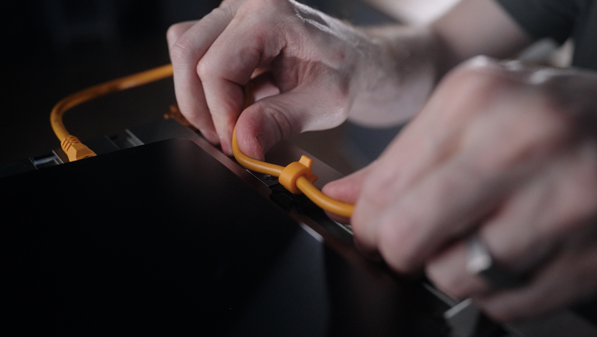 Two hands plug an orange Ethernet cable into a dark electronic device, close-up detail of the connection.",