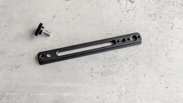 Black slender mounting plate with a long central slot and several holes, shown with a small screw knob nearby in the corner.