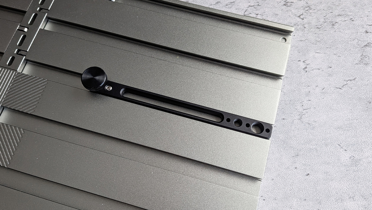 Close-up of a metallic camera slider rail with a black knurled adjustment knob and elongated slot, mounted on a grey surface