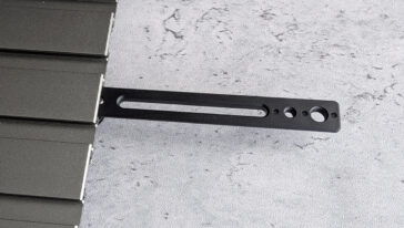 Black metal mounting bracket with a long oval slot and four circular holes, attached to the edge of a dark panel.