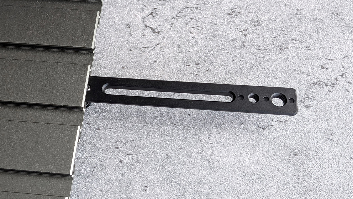 Black metal mounting bracket with a long oval slot and four circular holes, attached to the edge of a dark panel.