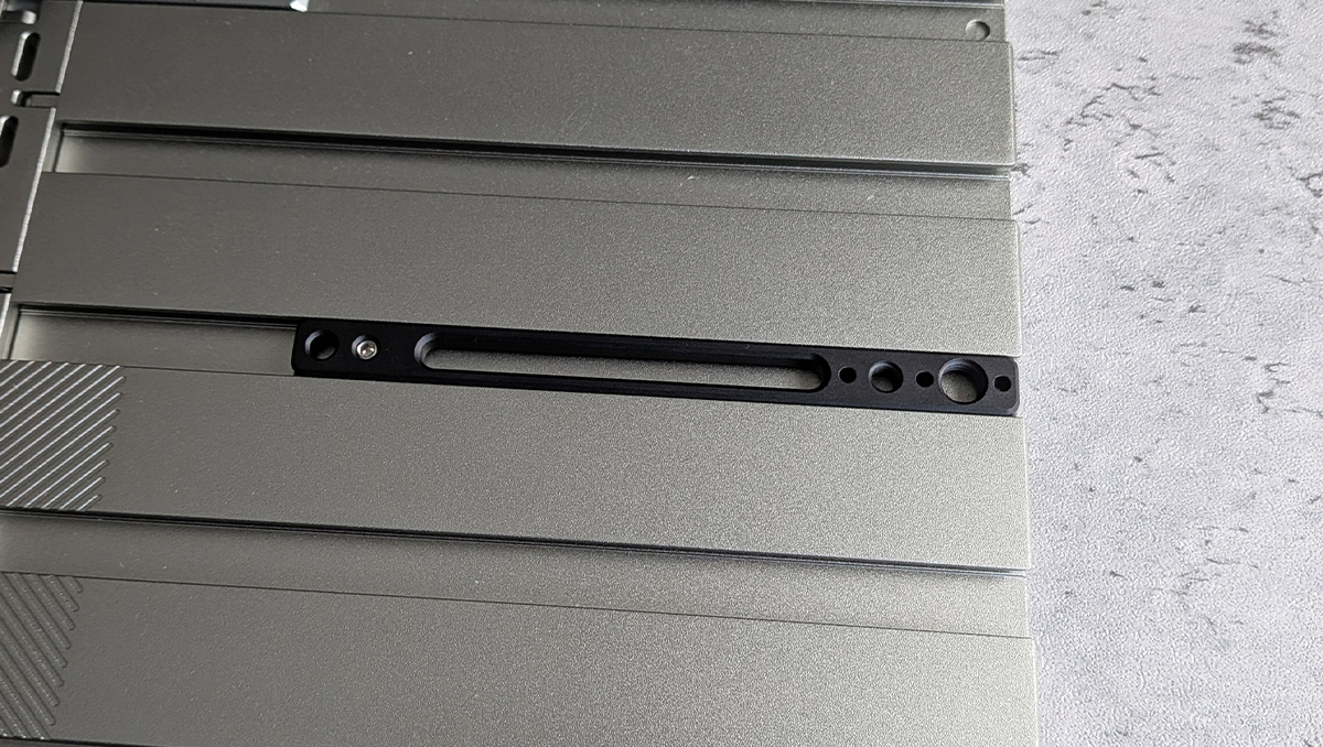Close-up of a gray metal panel featuring a long black carriage/slot with small circular holes along the edge, likely a mounting or retention area on hardware.