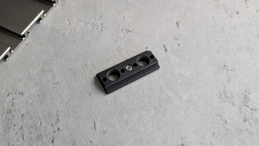 Small black plastic mounting block with three circular holes and a central screw on a gray textured surface