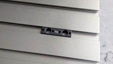 Black mounting bracket with two large holes and a central screw mounted on silver horizontal metal siding panels.