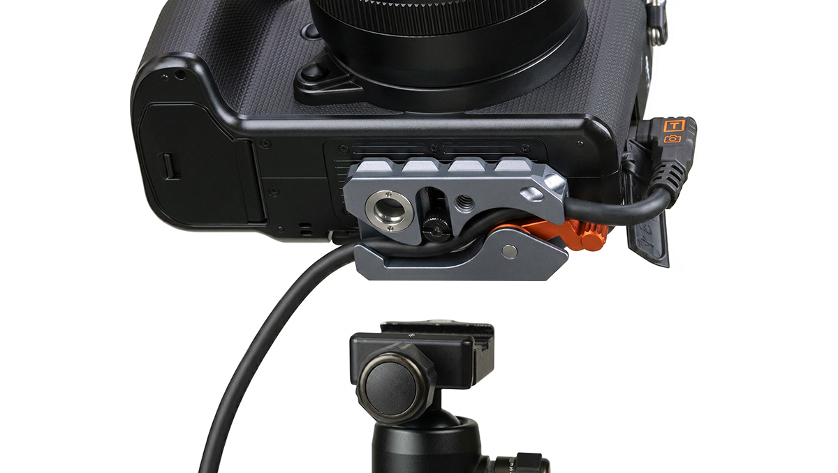 Close-up of a camera's bottom with a modular mounting rig and orange quick-release lever, connected by a cable to an accessory.