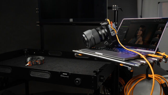 Camera tethered to a laptop on a wheeled desk in a dark studio, with orange cables and a strap nearby, ready for shoots or editing.