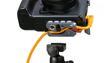 Close-up of a DSLR camera mounted on a tripod with an orange external power cable connected to a battery/dummy battery adapter on the side of the camera.