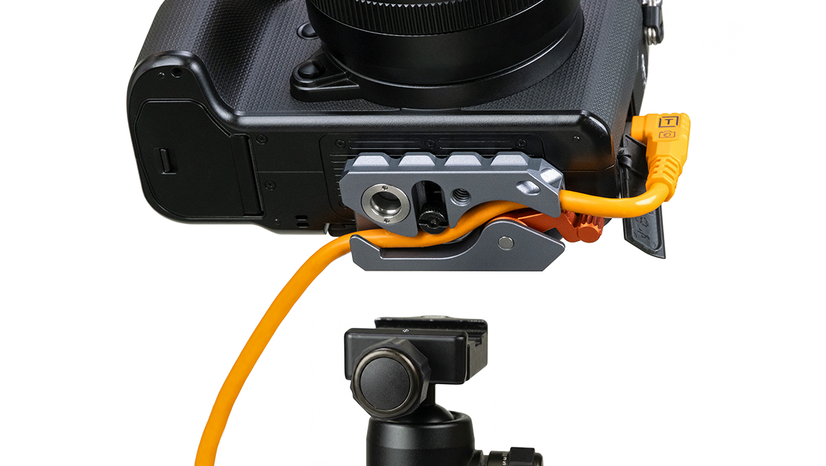 Close-up of a DSLR camera mounted on a tripod with an orange external power cable connected to a battery/dummy battery adapter on the side of the camera.